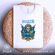Футболка водій військовий, біла, складена на плетеному кошику, фон білий, принт з черепом у військовій касці, двома автоматами та гербом України, текст 'ВОДІЙ' зверху принта.