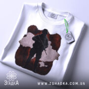 Футболка воїн України, біла, складена на столі, фон світлий, принт з силуетом солдата на червоному фоні з контуром України, з етикеткою на шнурку.