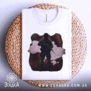 Футболка воїн України, біла, складена на плетеній кошику, фон світлий, принт з силуетом солдата на червоному фоні з контуром України.