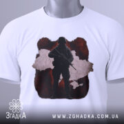 Футболка воїн України, біла, складена на столі, фон світлий, принт з силуетом солдата на червоному фоні з контуром України, з етикеткою на шнурку.