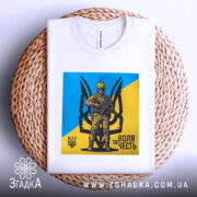 Футболка воля честь, біла, зображена на плетеній кошику, фон світло-сірий, принт з жовто-блакитним фоном та зображенням персонажа в військовій формі.