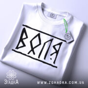 Футболка воля білого кольору, складена на поверхні, з чорним принтом 'ВОЛЯ', видно етикетку з зеленим шнурком, фон світло-сірий, ракурс зверху.