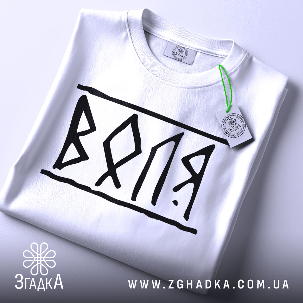 Футболка воля білого кольору, складена на поверхні, з чорним принтом 'ВОЛЯ', видно етикетку з зеленим шнурком, фон світло-сірий, ракурс зверху.