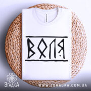 Футболка воля білого кольору, складена на плетеній кошику, з чорним принтом 'ВОЛЯ', фон світло-сірий, ракурс зверху.