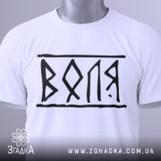 Футболка воля білого кольору, складена на поверхні, з чорним принтом 'ВОЛЯ', видно етикетку з зеленим шнурком, фон світло-сірий, ракурс зверху.