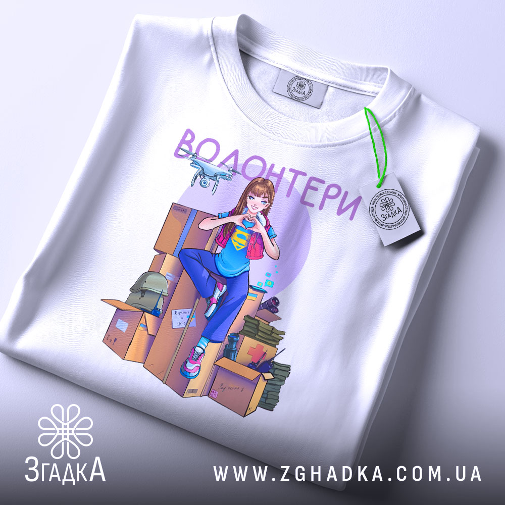Футболка волонтери білого кольору, складена на поверхні, ракурс зверху, фон світлий. Принт з дівчиною в синій футболці, яка робить жест серця, на фоні коробок і волонтерських предметів, з етикеткою на рукаві.