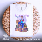 Футболка волонтери білого кольору, складена на плетеній основі, ракурс зверху, фон світлий. Принт з дівчиною в синій футболці, яка сидить на коробках, з елементами волонтерської діяльності, такими як дрон і медичні пакети.