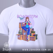 Футболка волонтери білого кольору, складена на поверхні, ракурс зверху, фон світлий. Принт з дівчиною в синій футболці, яка робить жест серця, на фоні коробок і волонтерських предметів, з етикеткою на рукаві.