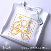 Футболка восьминіг у квадраті, біла, складена на столі, фон світло-сірий, принт з оранжевим восьминогом в квадраті, з етикеткою.