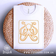 Футболка восьминіг у квадраті, біла, складена на кошику з плетеного матеріалу, фон світло-сірий, принт з оранжевим восьминогом в квадраті.