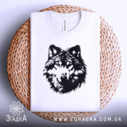 Футболка вовчий погляд, біла, складена на плоскій поверхні з плетеного кошика, фон світло-сірий, принт чорного кольору з зображенням голови вовка, видно етикетку на комірці.
