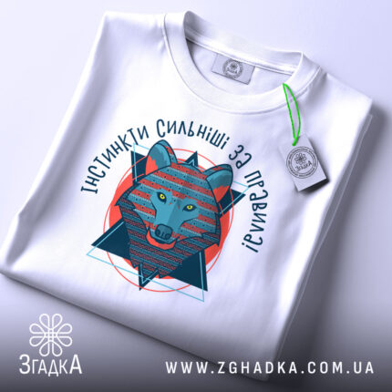 Футболка вовк інстинкти, складена біла футболка з принтом, ракурс зверху, фон світло-сірий, принт зображає вовка в геометричному стилі, текст 'ІНСТИНКТИ СИЛЬНІШІ ЗА ПРАВИЛА!' з яскравими кольорами.