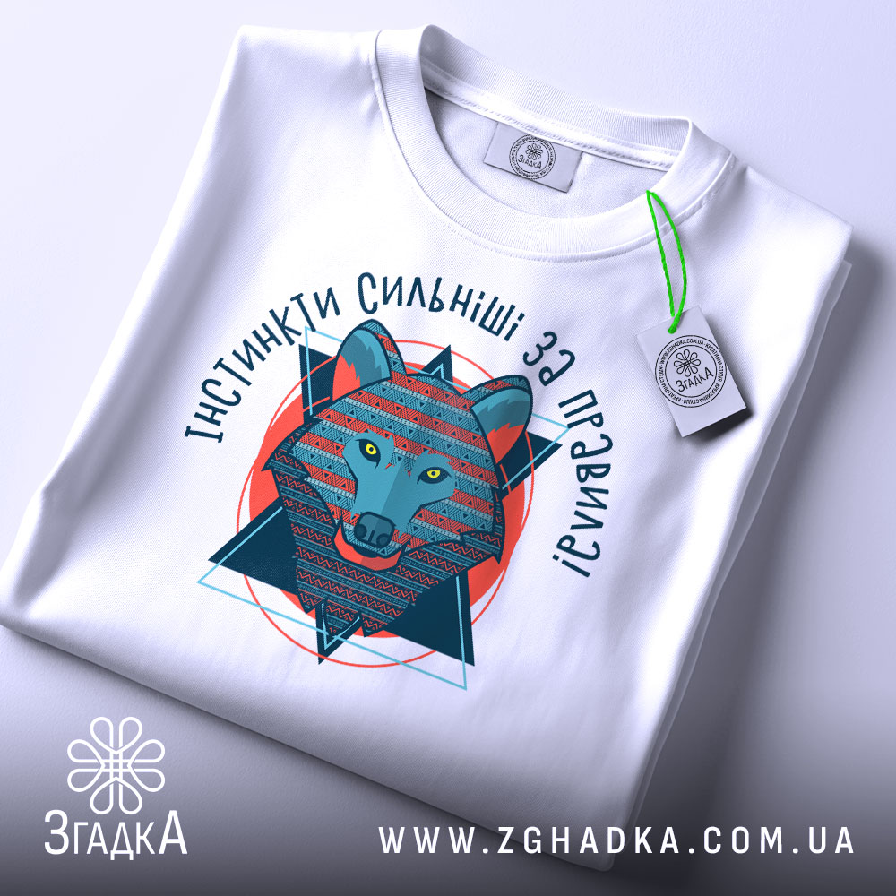 Футболка вовк інстинкти, складена біла футболка з принтом, ракурс зверху, фон світло-сірий, принт зображає вовка в геометричному стилі, текст 'ІНСТИНКТИ СИЛЬНІШІ ЗА ПРАВИЛА!' з яскравими кольорами.