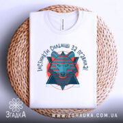 Футболка вовк інстинкти, складена біла футболка на плетеній кошику, ракурс зверху, фон світлий, принт з вовком в геометричному стилі, текст 'ІНСТИНКТИ СИЛЬНІШІ ЗА ПРАВИЛА!' у яскравих кольорах.