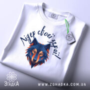 Футболка вовк лідер зграї, біла, складена на плоскій поверхні, фон світло-сірий, принт з геометричним зображенням вовка в синіх та оранжевих тонах, текст зверху написаний рукописним шрифтом, на футболці є етикетка з зеленим шнурком.