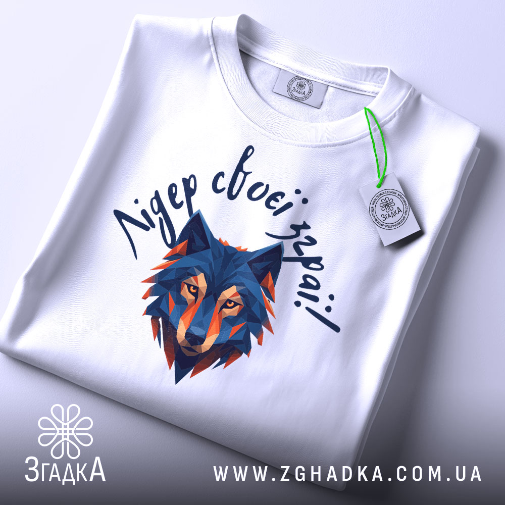 Футболка вовк лідер зграї, біла, складена на плоскій поверхні, фон світло-сірий, принт з геометричним зображенням вовка в синіх та оранжевих тонах, текст зверху написаний рукописним шрифтом, на футболці є етикетка з зеленим шнурком.