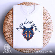 Футболка вовк лідер зграї, біла, складена на плоскій поверхні, фон світло-сірий, принт з геометричним зображенням вовка в синіх та оранжевих тонах, текст зверху написаний рукописним шрифтом, футболка лежить на плетеній основі.