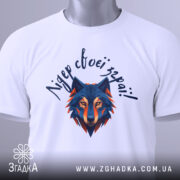 Футболка вовк лідер зграї, біла, складена на плоскій поверхні, фон світло-сірий, принт з геометричним зображенням вовка в синіх та оранжевих тонах, текст зверху написаний рукописним шрифтом, на футболці є етикетка з зеленим шнурком.