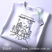 Футболка вовк лісовий господар, біла, складена на поверхні, зображена з верхнього ракурсу, фон світло-сірий, принт з чорними лініями, що зображує вовка на фоні гір і дерев, з етикеткою з зеленим шнурком.