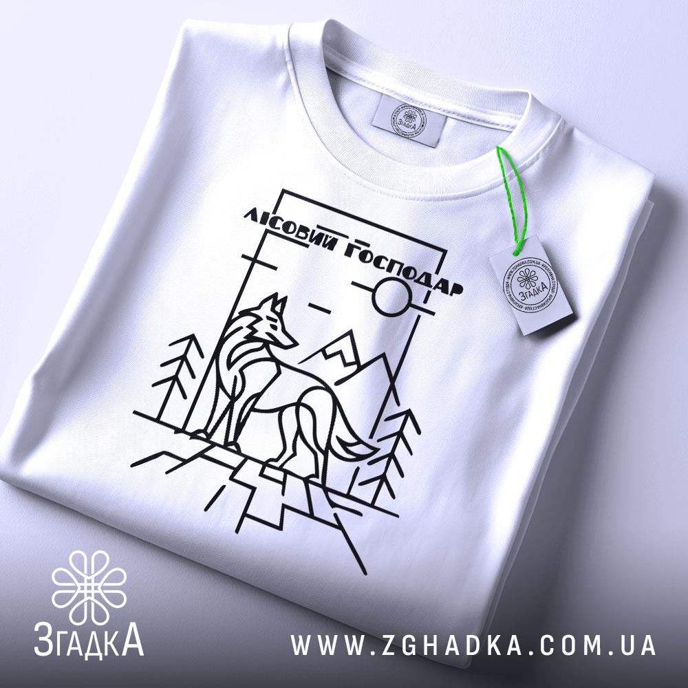 Футболка вовк лісовий господар, біла, складена на поверхні, зображена з верхнього ракурсу, фон світло-сірий, принт з чорними лініями, що зображує вовка на фоні гір і дерев, з етикеткою з зеленим шнурком.