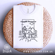 Футболка вовк лісовий господар, біла, складена на плетеній кошику, зображена з верхнього ракурсу, фон світло-сірий, принт з чорними лініями, що зображує вовка на фоні гір і дерев.