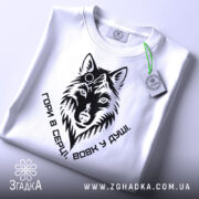 Футболка вовк у душі, біла, складена на столі, фон світлий, принт чорного кольору з зображенням вовка та написом 'Гори в серці, вовк у душі', на футболці є етикетка з зеленим шнурком.