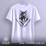 Футболка вовк у душі, біла, підвішена на вішалці, фон світлий, принт чорного кольору з зображенням вовка та написом 'Гори в серці, вовк у душі'.