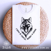 Футболка вовк у душі, біла, складена на плетеній кошику, фон світлий, принт чорного кольору з зображенням вовка та написом 'Гори в серці, вовк у душі'.