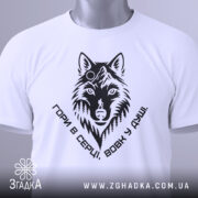 Футболка вовк у душі, біла, складена на столі, фон світлий, принт чорного кольору з зображенням вовка та написом 'Гори в серці, вовк у душі', на футболці є етикетка з зеленим шнурком.