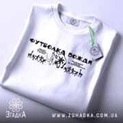 Футболка вождя нашого білого кольору, складена на поверхні, з принтом чорного кольору, з етикеткою зеленого кольору, на фоні світло-сірого градієнту, з чітким зображенням фігурок людей та слона.