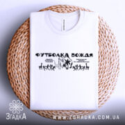 Футболка вождя нашого білого кольору, складена на плетеній кошику, з принтом чорного кольору, на фоні світло-сірого градієнту, з чітким зображенням фігурок людей та пальм.