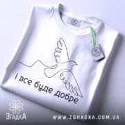 Футболка все буде добре. Біла футболка складена на столі, з видимим етикеткою. Фон світлий, з легким градієнтом. Принт з птахом та написом 'і все буде добре' розташований по центру.