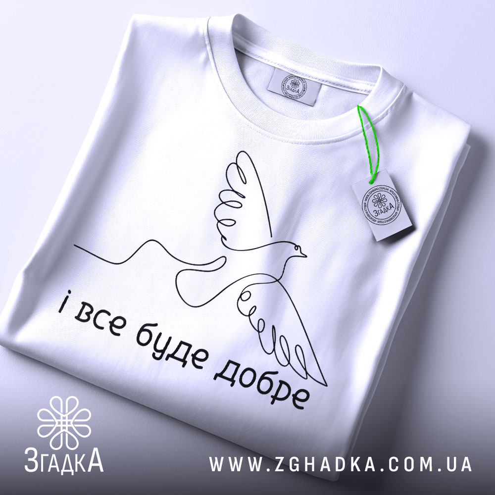 Футболка все буде добре. Біла футболка складена на столі, з видимим етикеткою. Фон світлий, з легким градієнтом. Принт з птахом та написом 'і все буде добре' розташований по центру.