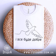 Футболка все буде добре. Біла футболка складена на плетеній кошику, фон світлий. Принт з птахом та написом 'і все буде добре' розташований по центру.