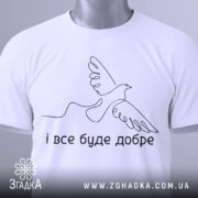 Футболка все буде добре. Біла футболка складена на столі, з видимим етикеткою. Фон світлий, з легким градієнтом. Принт з птахом та написом 'і все буде добре' розташований по центру.
