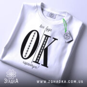 Футболка все буде ок, білого кольору, складена на поверхні, зображена з верхнього ракурсу, фон світлий, принт чорного кольору з великими літерами 'OK' та текстом 'все буде гарантію!' у верхній частині, на футболці є етикетка з зеленим шнурком.