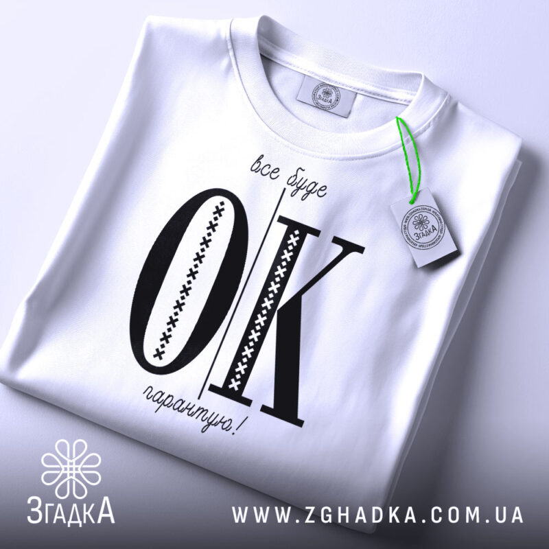 Футболка все буде ок, білого кольору, складена на поверхні, зображена з верхнього ракурсу, фон світлий, принт чорного кольору з великими літерами 'OK' та текстом 'все буде гарантію!' у верхній частині, на футболці є етикетка з зеленим шнурком.