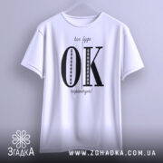 Футболка все буде ок, білого кольору, зображена з фронтального ракурсу на вішалці, фон світлий, принт чорного кольору з великими літерами 'OK' та текстом 'все буде гарантію!' у верхній частині.