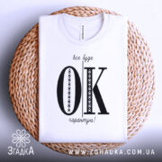 Футболка все буде ок, білого кольору, складена на плетеній основі, зображена з верхнього ракурсу, фон світлий, принт чорного кольору з великими літерами 'OK' та текстом 'все буде гарантію!' у верхній частині.
