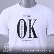 Футболка все буде ок, білого кольору, складена на поверхні, зображена з верхнього ракурсу, фон світлий, принт чорного кольору з великими літерами 'OK' та текстом 'все буде гарантію!' у верхній частині, на футболці є етикетка з зеленим шнурком.