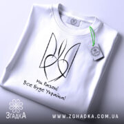 Футболка все буде Україна, біла, складена на столі, фон світло-сірий, принт чорного кольору з написом 'Ми Разом! Все буде Україна!' та стилізованим гербом, з етикеткою зеленого кольору.