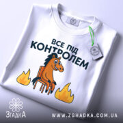 Футболка все під контролем, білого кольору, складена на поверхні, фон світлий, принт з конем на стільці та вогнем, прикріплена етикетка з зеленим шнурком.