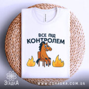 Футболка все під контролем, білого кольору, складена на плетеній основі, фон світлий, принт з конем на стільці та вогнем, видно деталі комірця.