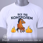Футболка все під контролем, білого кольору, складена на поверхні, фон світлий, принт з конем на стільці та вогнем, прикріплена етикетка з зеленим шнурком.