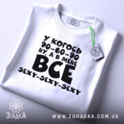 Футболка все сексі вайб, складена біла футболка з чорним принтом, зображена зверху, фон світлий, на футболці є етикетка з зеленим шнурком, принт містить текст з жартом про розміри.