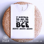 Футболка все сексі вайб, складена біла футболка з чорним принтом, зображена зверху на плетеній основі, фон світлий, принт містить текст з жартом про розміри.