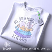 Футболка все що не робиться, білого кольору, зображена зверху, на фоні світло-сірого градієнту. Принт з котом на кольорових паперах, з зеленим ярликом, що підкреслює стиль.