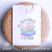 Футболка все що не робиться, білого кольору, розкладена на плетеній кошику, фон світло-сірий. Принт з котом на кольорових паперах, текст українською мовою.