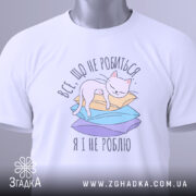 Футболка все що не робиться, білого кольору, зображена зверху, на фоні світло-сірого градієнту. Принт з котом на кольорових паперах, з зеленим ярликом, що підкреслює стиль.