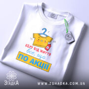 Футболка все, що по акції. Біла футболка, складена на столі, з видимим принтом. Фон світло-сірий, принт кольоровий, з жовтою футболкою на вішалці та червоною пляшечкою, текст червоний і жовтий, з зеленим ярликом.